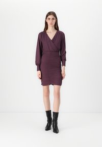 Violetinė suknelė su ilgomis pūstomis rankovėmis ir giliu V formos iškirpimu. Suknelė turi raukšlėjimą per visą kūną, baigiasi virš kelių, derinama su juodais batais.