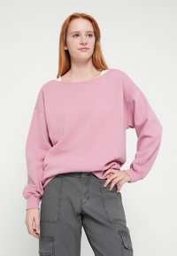 Rózsaszín pulóver, laza szabással, széles nyakkivágással és hosszú ujjakkal, szürke cargo nadrággal párosítva, oldal zsebekkel és texturált anyaggal.