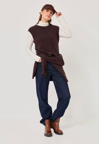 Brown sleeveless knit tunic over a white turtleneck, paired with loose blue jeans and brown ankle boots. The outfit includes a matching cap.
