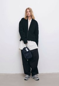 Mustaa ribattua vetoketjullista takkia valkoisen kauluspaidan päällä, yhdistettynä väljään mustaan housuun ja harmaisiin lenkkareihin. Pitelee pientä mustaa laukkua.