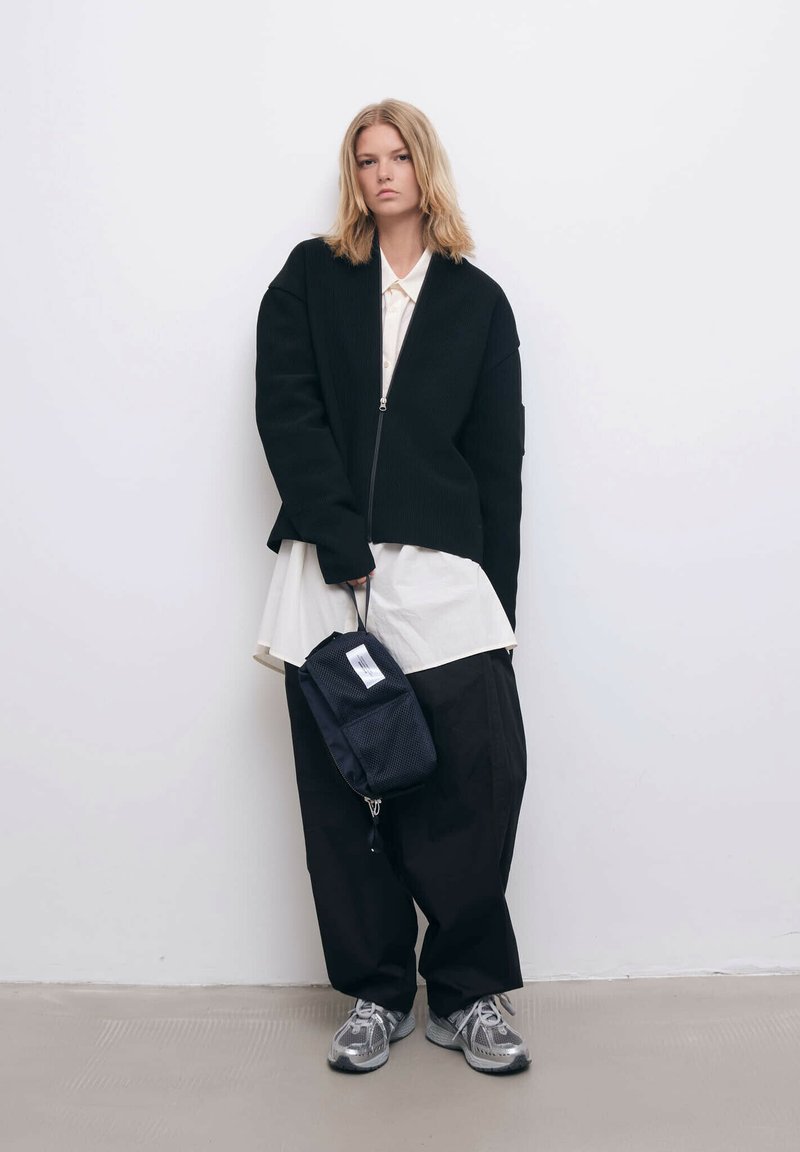 Mustaa ribattua vetoketjullista takkia valkoisen kauluspaidan päällä, yhdistettynä väljään mustaan housuun ja harmaisiin lenkkareihin. Pitelee pientä mustaa laukkua.