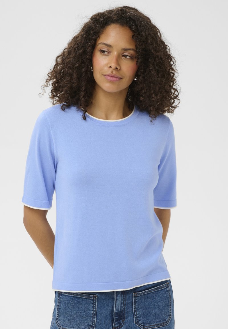 Femme aux cheveux bouclés portant un haut à manches courtes bleu clair avec des bordures blanches et un jean bleu, se tenant devant un fond uni.