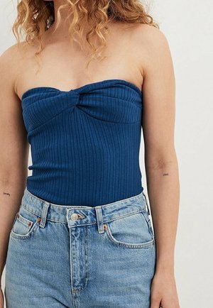 Kvinde iført en blå stropfri ribbet top kombineret med højtaljede lyseblå jeans, stående mod en ensfarvet baggrund.