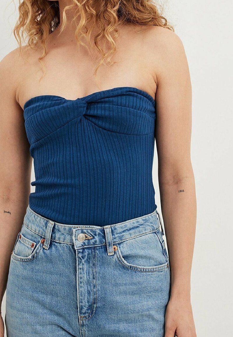 Femme portant un haut bleu sans bretelles côtelé associé à un jean taille haute bleu clair, debout sur un fond uni.