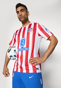 Červeno-biely pruhovaný futbalový dres s modrými akcentmi, s klubovým logom, grafikou sponzora a krátkymi rukávmi. Hráč drží futbalovú loptu.