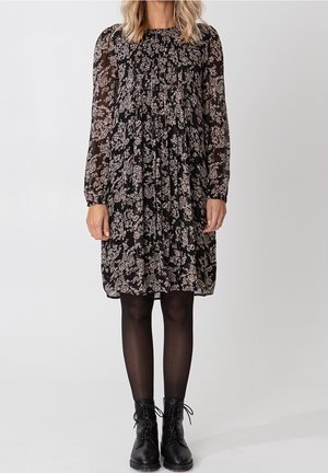 Vrouw draagt een zwarte knielange jurk met wit bloemenpatroon, lange doorzichtige mouwen, zwarte panty's en zwarte veter enkellaarzen.