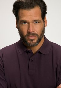 Dunkellila Polo-Shirt mit einer Drei-Knopf-Leiste, subtiler Textur und einem klassischen Kragen-Design. Männliches Model mit vollem Bart und kurzen Haaren.