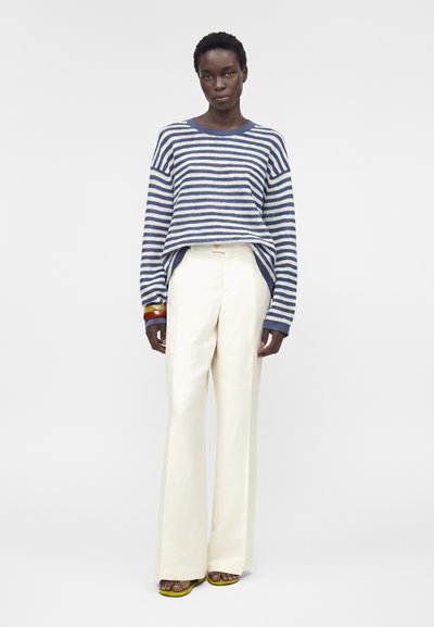 Γυναίκα όρθια που φοράει μπλε και άσπρη ριγέ μπλούζα, λευκά παντελόνια με φαρδιά πόδια, κίτρινες σανδάλες και πολύχρωμα βραχιόλια στον καρπό της.