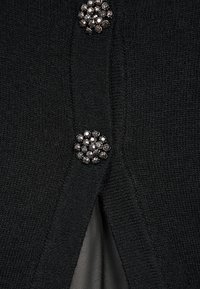 Czarny dzianinowy kardigan z fakturowanej tkaniny, poziomym rozcięciem oraz grupą dekoracyjnych guzików z kryształkami przy zapięciu.