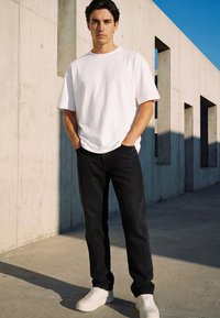 Young man stands with hands in pockets wearing white t-shirt, black jeans, and white sneakers against a concrete wall under clear sky.