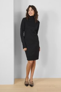 Black knit turtleneck dress with long sleeves and ribbed hem. Paired with pointed-toe black heels featuring strap accents. Simple and sleek design.