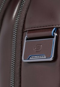 Dettaglio di una borsa in pelle bordeaux con una cerniera metallica argentata e una targhetta rettangolare blu con il logo "Piquadro".