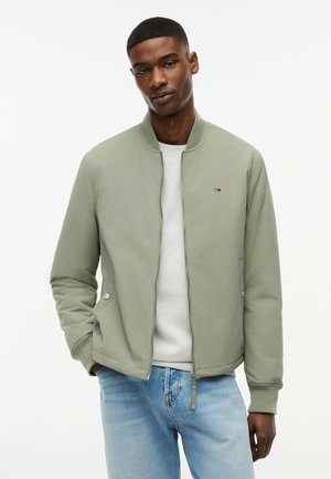 Mann trägt eine hellgrüne Jacke mit Reißverschluss über einem weißen Hemd und hellblauen Jeans, steht mit den Händen in den Taschen vor weißem Hintergrund.