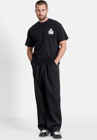 Black cotton t-shirt with a small white graphic on the chest, paired with loose black trousers and black shoes with white accents.