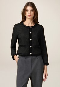 Chaqueta de tweed negra con cuello redondo, bolsillos frontales y cinco grandes botones plateados. Presenta una tela texturizada y un diseño cropped.