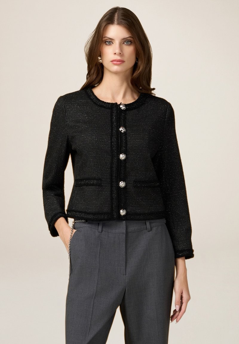 Chaqueta de tweed negra con cuello redondo, bolsillos frontales y cinco grandes botones plateados. Presenta una tela texturizada y un diseño cropped.