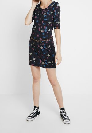 Femme portant une robe courte à fleurs sombres avec une ceinture tressée et des baskets montantes noires, se tenant devant un fond blanc uni.