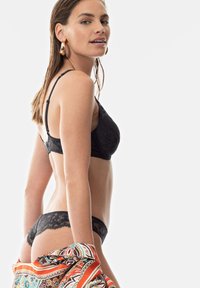 Reggiseno in pizzo nero e perizoma abbinato, caratterizzati da delicati motivi floreali. Il modello tiene una sciarpa colorata e decorata, mettendo in risalto curve e texture.