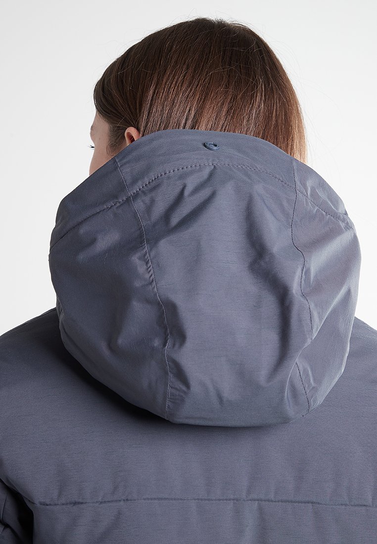 Persona con cabello castaño claro, vistiendo una chaqueta gris con capucha adjunta, vista desde atrás sobre un fondo blanco liso.