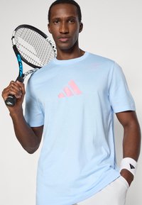 Jasnoniebieska koszulka sportowa z krótkim rękawem, z różowym logo Adidas. Model trzyma czarno-niebieską rakietę tenisową i ma na sobie białą opaskę na nadgarstek.