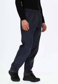 Marineblaue wasserdichte Hose mit elastischem Bund, Seitentaschen und tailliertem Schnitt. Kombiniert mit schwarzen geschnürten Schuhen mit einer robusten Sohle.