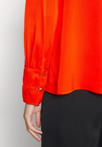 Blouse orange vif avec des poignets côtelés et des détails de boutons. Tissu lisse et brillant avec une coupe décontractée, accompagné de pantalons noirs.