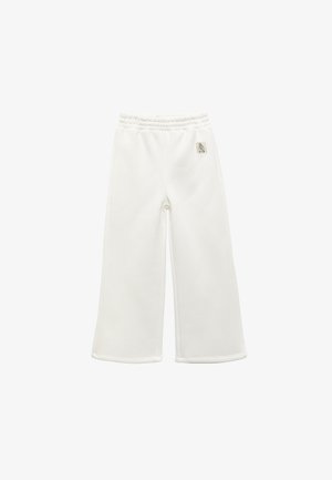 Valkoiset leveälahkeiset housut, joissa on elastinen vyötärö ja pieni logo-patch oikealla lantiolla, asetettuna tasaisesti valkoista taustaa vasten.