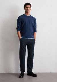 Granatowy sweter z jasnoniebieskim kołnierzem, zestawiony z ciemnymi spodniami i czarnymi butami. Model stoi z rękami w kieszeniach, na neutralnym tle.