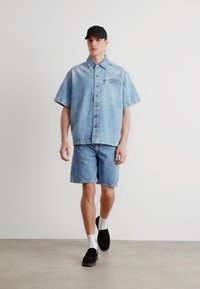 Camisa de mezclilla de manga corta azul claro con bolsillo frontal, combinada con pantalones cortos de mezclilla a juego. Gorra negra, calcetines blancos y zapatos negros sin cordones.