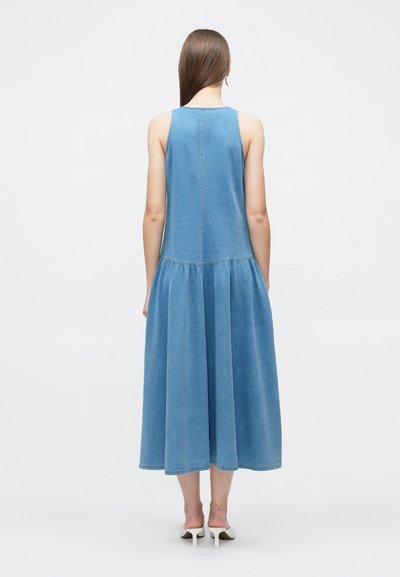 Γυναίκα με ίσια καστανά μαλλιά φορώντας αμάνικο μπλε τζιν φόρεμα μέσης και λευκές πέδιλες με τακούνι, όρθια με την πλάτη στην κάμερα.