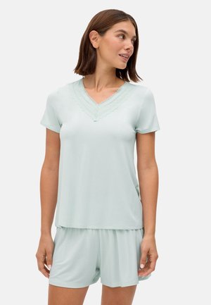 Mujer con blusa de manga corta y escote en V de color azul pálido con borde de encaje y pantalones cortos sueltos a juego, de pie contra un fondo liso y claro.