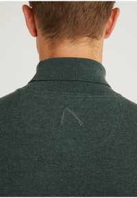 Dunkelgrüner Pullover mit geripptem Kragen und einem kleinen sichtbaren Faden auf der Rückseite. Weiche Textur und figurbetontes Design.