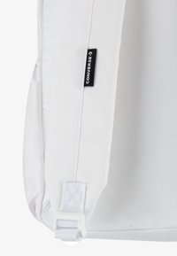 Vit kanvasryggsäck med en svart "CONVERSE"-etikett, som har en enkel design, rektangulär form och robusta vita detaljer.