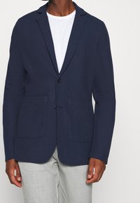 Navy blazer with a textured fabric, single-button closure, notched lapels, and two patch pockets, worn over a white shirt.