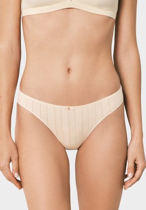 Femme portant un sous-vêtement style bikini beige à motifs et un soutien-gorge assorti sur fond uni, torse visible de la poitrine aux cuisses supérieures.