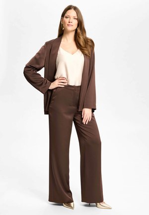 Femme aux longs cheveux portant des boucles d’oreilles créoles en or, une blouse crème, un blazer marron et un pantalon large, ainsi que des talons dorés à bout pointu, debout avec assurance.