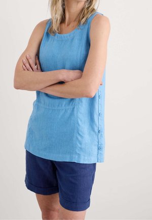 Vrouw met gekruiste armen, gekleed in een mouwloos lichtblauw topje met knopen aan de zijkant en donkerblauwe opgerolde shorts.