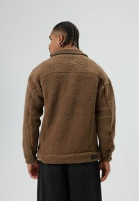 Brown fleece jacket featuring a textured exterior, large collar, and buttoned cuffs. Label visible at the hem, styled with black pants.