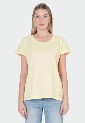 Vrouw draagt een lichtgeel T-shirt met korte mouwen en lichtblauwe spijkerbroek, staand tegen een effen lichtgrijze achtergrond.