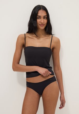 Donna con capelli lunghi e neri che indossa un top nero con spalline sottili e biancheria intima nera abbinata con rifiniture in pizzo, in piedi davanti a uno sfondo chiaro e semplice.