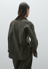 Chemise vert foncé, ample, avec un col, des poignets à boutons et une texture lisse. Présente un ourlet arrondi et un design minimaliste.