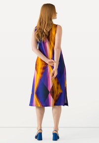 Ein ärmelloses, knielanges Kleid mit geometrischen Mustern in lila, orange und pink. Das Model trägt blaue Blockabsatzschuhe.