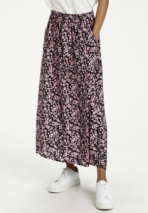 Person trägt einen schwarzen und rosa Maxirock mit Blumenmuster und Taschen sowie weiße Sneakers, steht vor einem einfarbigen Hintergrund.