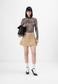 Vrouw in een leopardprint langemouwtop, beige rok, witte sokken, zwarte hakken, met een grote zwarte clutch tegen een witte achtergrond.