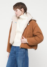 Jachetă insulată maro cu căptușeală din fleece bej, având glugă, fermoar frontal și două buzunare laterale. Se poartă peste un pulover alb din tricot.