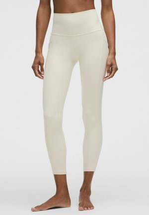 Kvinde iført højtsiddende, cremefarvede tætsiddende leggings, stående barfodet på en ensartet lys baggrund.