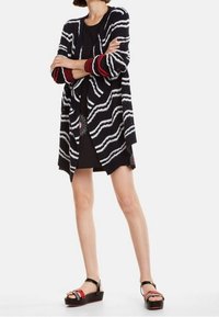 Cardigan a strisce bianche e nere con dettagli rossi, apertura frontale, maniche lunghe e vestibilità comoda, indossato sopra un vestito nero e sandali con zeppa.