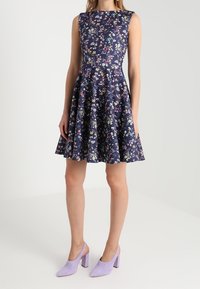 Knytklänning utan ärmar med blommigt tryck i marinblått och en utsvängd kjol. Texturerat tyg med små blommor i flera färger. Matchas med lila klackskor.