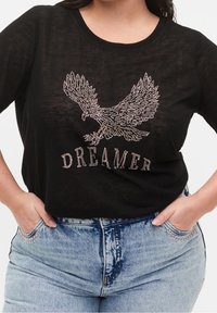 Černé triko s dlouhým rukávem má lesklý design orla vyrobený z rhinestones nad slovem "DREAMER" a je kombinováno s světle modrými potrhanými džíny.