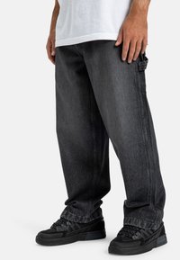 Ciemnoszare dżinsowe spodnie cargo o luźnym kroju, z bocznymi kieszeniami i widocznymi szwami. W zestawieniu z czarnymi butami sportowymi.